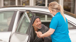Female caregiver helps elderly patient out of the back seat of a car.
